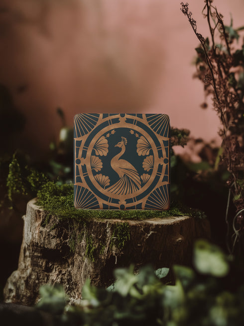 Square ceramic coaster featuring an Art Deco copper peacock design centered in a medallion, surrounded by fan patterns and shell motifs on a dark forest green background, with cork backing.